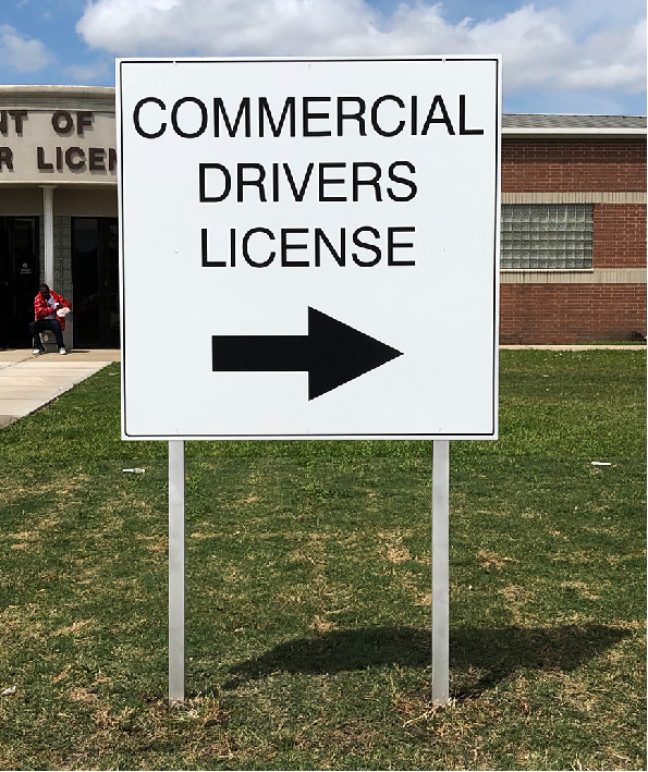 A sign reads "Commercial Drivers License" with an arrow pointing right, positioned on grass near a brick building and a sidewalk.