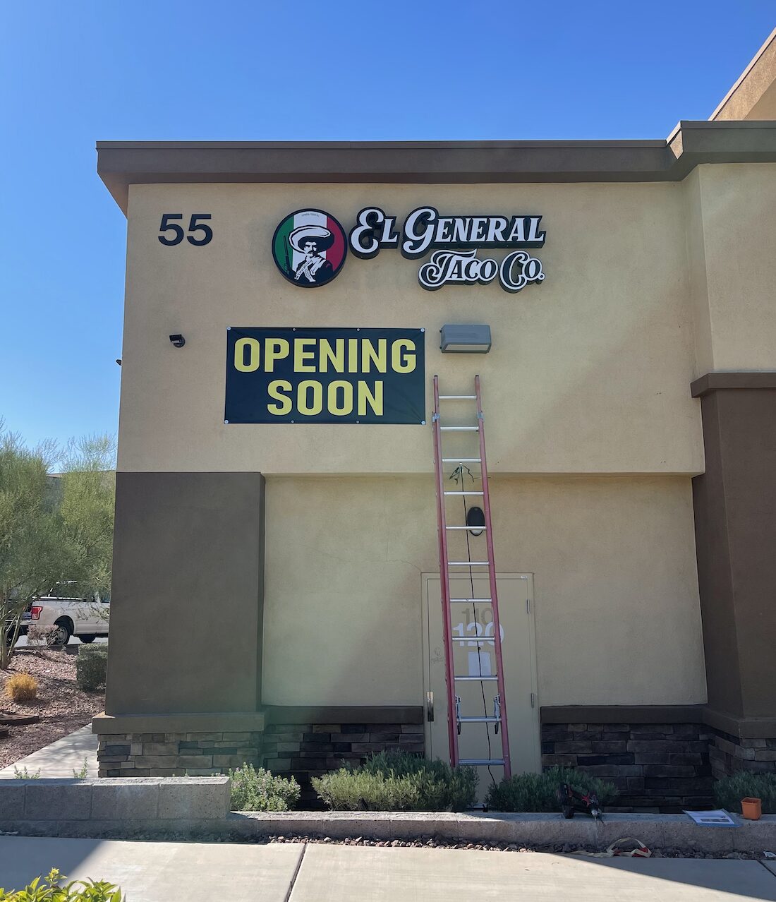 A building with a sign that reads "El General Taco Co." and a banner below it stating "OPENING SOON." A ladder is propped up against the building. The number "55" is visible on the wall.