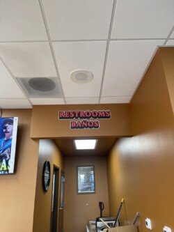Restroom entrance with a sign reading "RESTROOMS BAÑOS" in a hallway with brown walls and ceiling tiles. A cleaning cart is seen to the right.