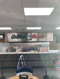 A restaurant counter with a sign above it reading "Grilled Fresh." The counter includes drink dispensers, condiment containers, and a stack of trays.