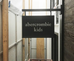 A black sign with white text reads "abercrombie kids," displayed in a narrow passageway under construction scaffolding.