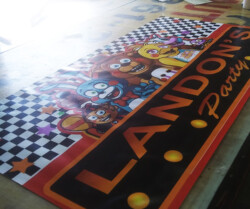 A colorful banner with cartoon characters, reading "Landon's Party" in bold letters on a table. The design features a checkered pattern and stars.