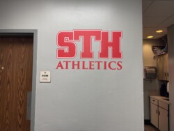 Wall with "STH ATHLETICS" in red letters, a door with a sign labeled "GYM OFFICE Director of Athletics," and an office corner with cabinets and a paper towel dispenser.