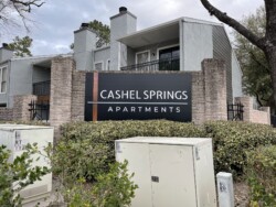 A sign reading "Cashel Springs Apartments" is displayed in front of a gray, multi-unit residential building with brick pillars and greenery around. Electrical utility boxes are visible in the foreground.