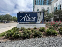 Sign for "Allure Hermann Park Luxury Apartments" with buildings and landscaped greenery in the background.