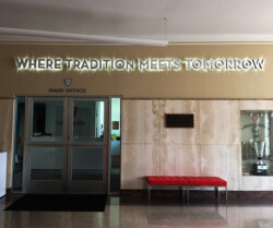 Entrance to a main office with a sign above reading "Where Tradition Meets Tomorrow." The area features a glass door, a red bench on the right, and a trophy case in the right side background.