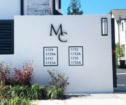A white wall with the letters "MC" and addresses 1729, 1729A, 1731, 1731A, 1733, 1733A, 1735, 1735A listed, with some plants in front and residential buildings in the background.