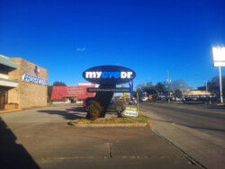 A commercial street featuring a "myeyedr" optometry sign. To the left is a Progressive building. A "For Lease" sign is on a patch of grass in front of the sidewalk. The sky is clear and blue.