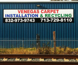 Sign on a blue building reads "Venegas Carpet Installation & Recycling" with two phone numbers: 832-973-9743 and 713-729-8110. A chain-link fence and railroad tracks are in the foreground.
