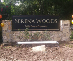 A stone sign reading "Serena Woods: A Villa Serena Community" with the number 2800 to the right, surrounded by trees and decorations.