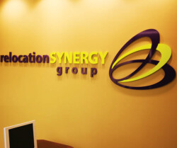 Office wall with a sign that reads "Relocation SYNERGY group" featuring a logo with intertwined shapes in purple and yellow, next to a computer monitor.