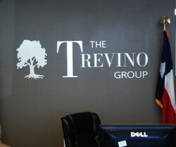 Office wall with "The Trevino Group" logo, a white tree graphic, and a Texas flag. A Dell monitor and chair are in the foreground.