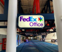 A FedEx Office sign hangs from the ceiling in an indoor corridor with a blue carpet.