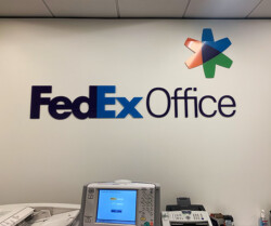 Interior of a FedEx Office location showing a wall with the FedEx Office logo and a counter with office equipment including a printer and a phone.