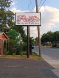 Sign reads "Puebla's Mexican Kitchen" on a tall signpost, positioned near a tree-lined street with houses and a power line in the background, under a partly cloudy sky.