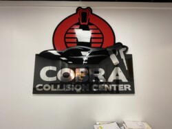 A wall-mounted sign reads "Cobra Collision Center" with an image of a car and a spray paint gun. A stylized cobra emblem is positioned above the text. Office documents are stacked on a counter below.