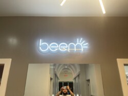 A person taking a selfie in front of a mirror with a neon sign that reads "beem" on the wall above.
