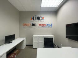 A small office with white furniture, a black chair, and logos for "LINC," "SMARTLINC," and "REDFIN Plumbing Co." on the wall. A flat-screen TV is mounted on the right wall.
