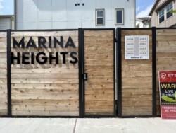 A wooden fence with a gate sign displaying "MARINA HEIGHTS" and building unit numbers. A warning and trespassing sign is on the right.
