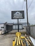 Sign for "Teejay Machine Co." with a contact number, located behind a metal gate with industrial pipes and equipment in the foreground. Overcast sky and utility poles in the background.