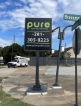 Sign for "Pure Auto Spa + Detailing" with a contact number and website at an intersection of Braesmain Drive. Visible are a truck, buildings, and utility poles under a partly cloudy sky.