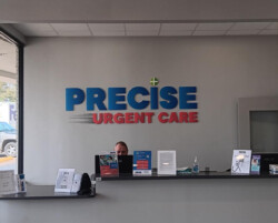 Front desk area of Precise Urgent Care with a staff member seated at a computer. Signs, brochures, and hand sanitizer are on the counter. The wall behind features the clinic’s name in blue and red letters.