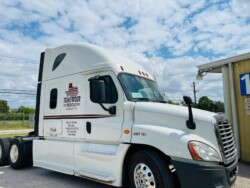 A white semi-truck with "Texas Pride Elite" and "UNIT 103" written on the side parked near a yellow building under a partly cloudy sky.