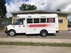 A white school bus with "St. Thomas High School" and website details on the side is parked on a paved surface with a yellow building in the background.
